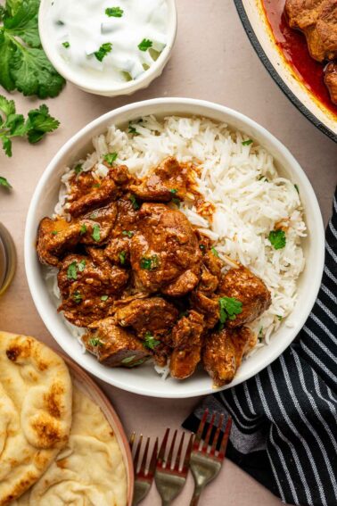 A bowl of white rice topped with rogan josh, garnished with cilantro, served with naan, yogurt, and fresh cilantro on the side.