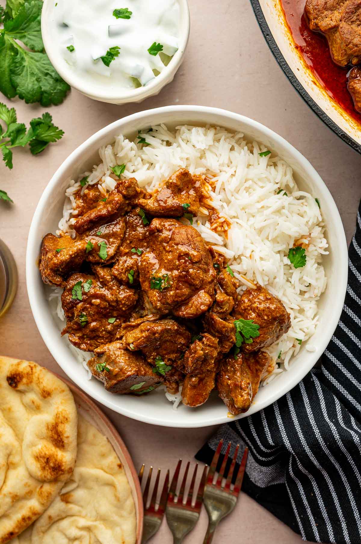 A bowl of white rice topped with rogan josh, garnished with cilantro, served with naan, yogurt, and fresh cilantro on the side.