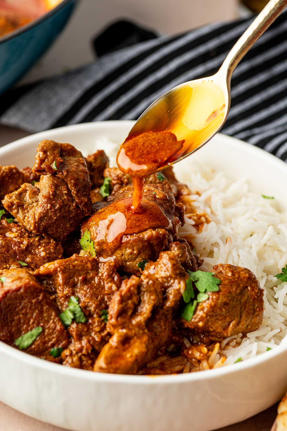 A bowl of white rice topped with chunks of rogan josh curry, garnished with herbs, with sauce being spooned over the curry.
