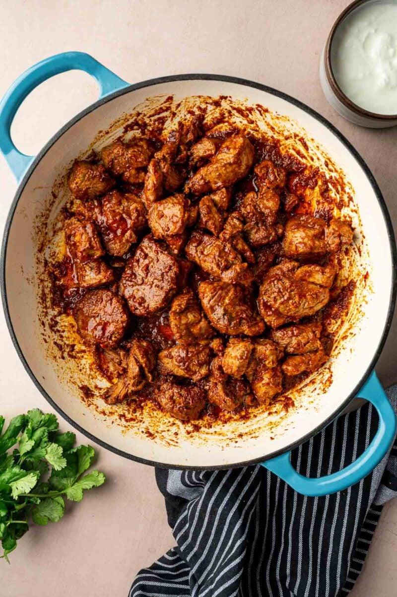 Chunks of cooked, seasoned lamb in a blue Dutch oven, surrounded by a bowl of yogurt, fresh cilantro, and a striped cloth.