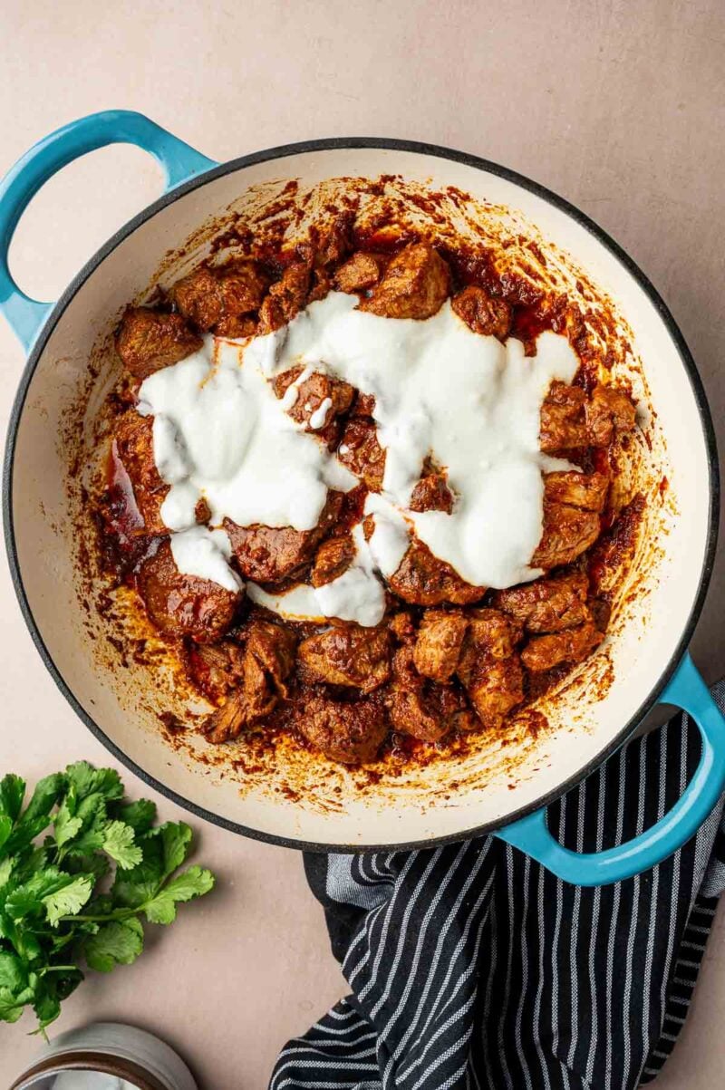 A blue Dutch oven filled with cooked pieces of lamb in a red curry sauce, topped with a swirl of white yogurt, sits on a beige surface next to a bunch of parsley and a striped towel.