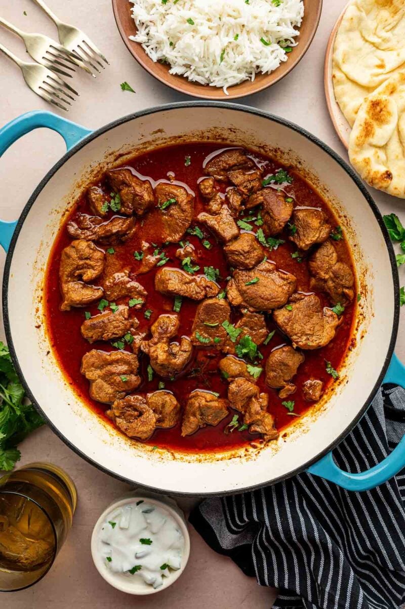 A pot of spicy rogan josh garnished with cilantro, surrounded by a plate of rice, naan bread, forks, a glass of water, and a bowl of raita sauce.