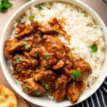 A bowl of Rogan Josh curry served with white rice and garnished with cilantro, next to naan bread and a fork on a striped napkin.