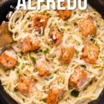 A skillet filled with creamy salmon Alfredo topped with chunks of cooked salmon, garnished with parsley and Parmesan cheese.