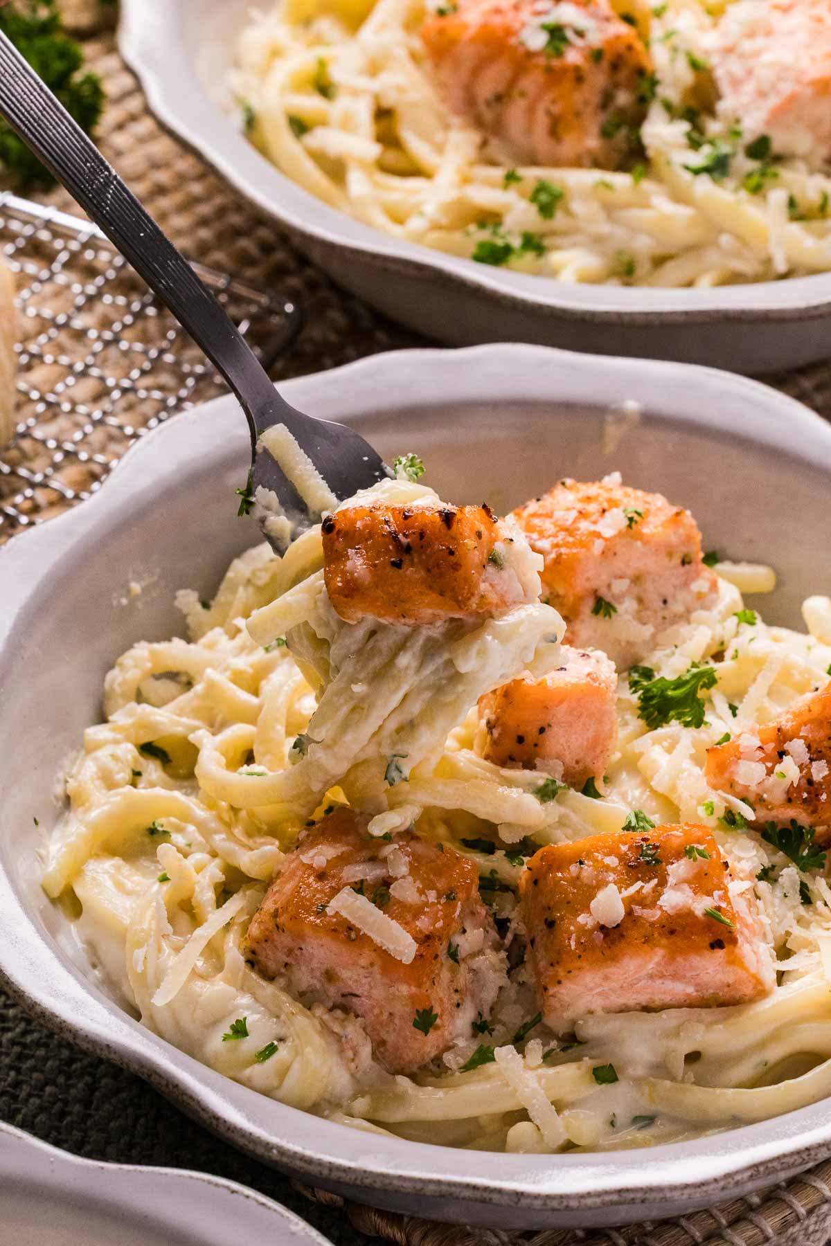 A bowl of salmon alfredo, garnished with grated cheese and parsley; a fork holds a bite of pasta and salmon.