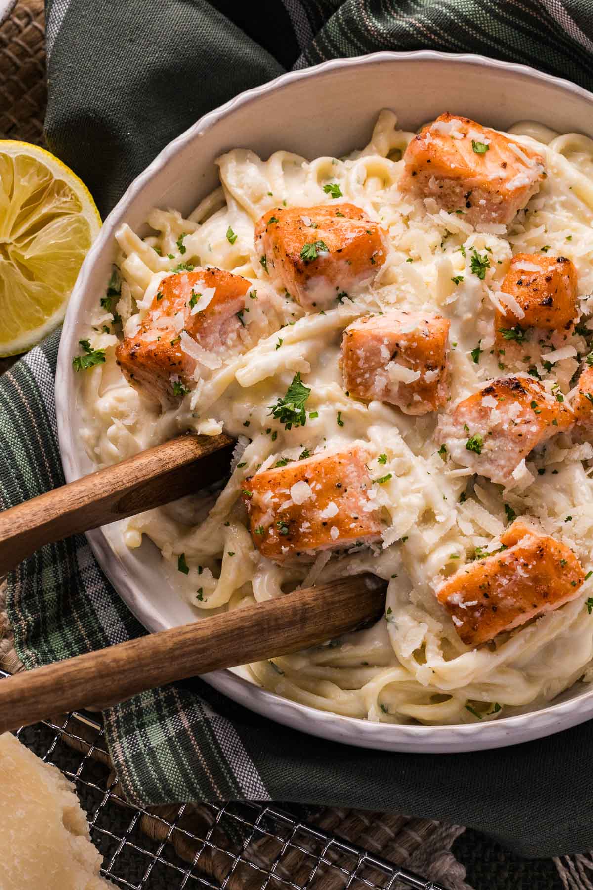 A bowl of linguini pasta topped with salmon pieces in alfredo sauce, garnished with parsley and grated cheese, with wooden tongs and a lemon half beside the dish.