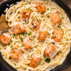A skillet filled with creamy alfredo pasta topped with seared salmon pieces, garnished with chopped parsley and grated cheese.