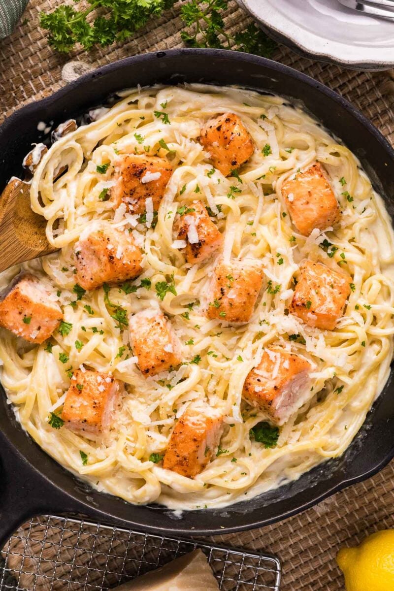 A skillet filled with creamy alfredo pasta topped with seared salmon pieces, garnished with chopped parsley and grated cheese.