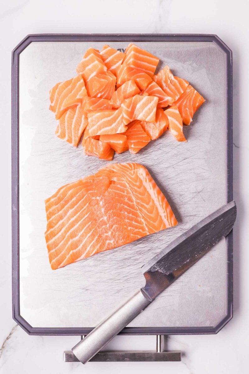 Raw salmon fillet and salmon cubes on a metal cutting board with a knife placed beside them.