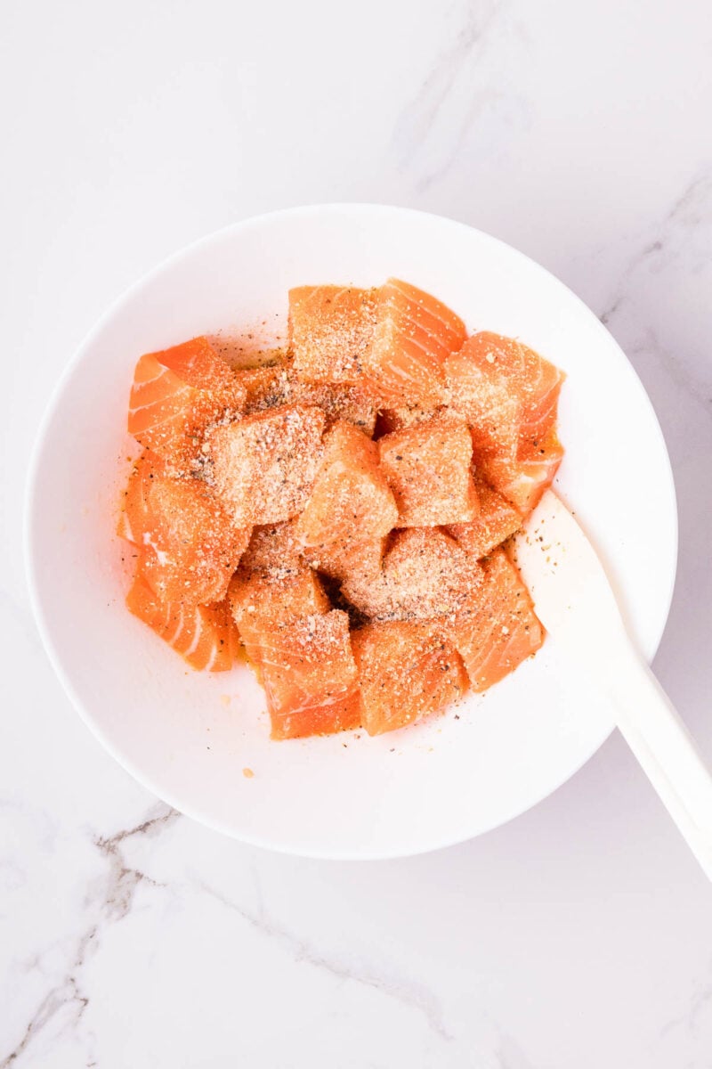 A white bowl contains raw salmon cubes sprinkled with seasoning, with a white spatula resting inside the bowl on a marble surface.
