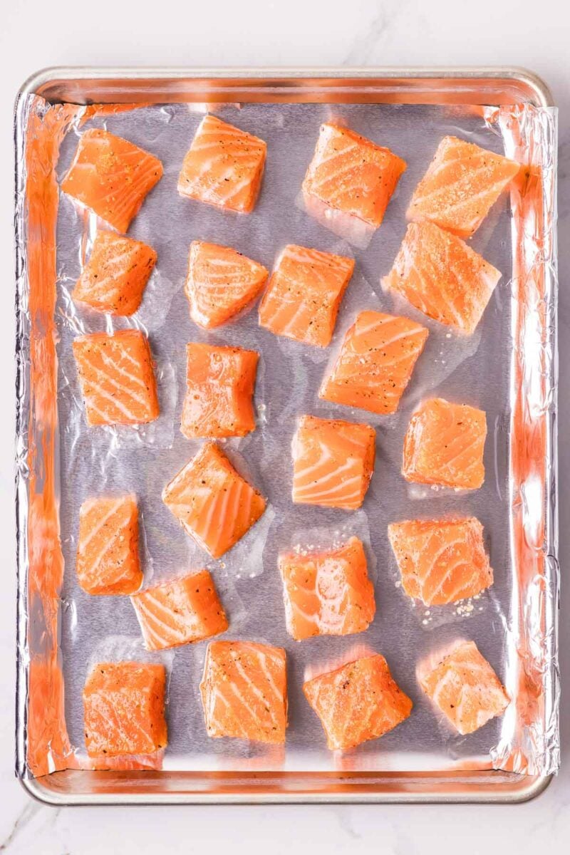 Twenty raw salmon cubes, seasoned with salt and pepper, are arranged evenly on a foil-lined baking sheet.