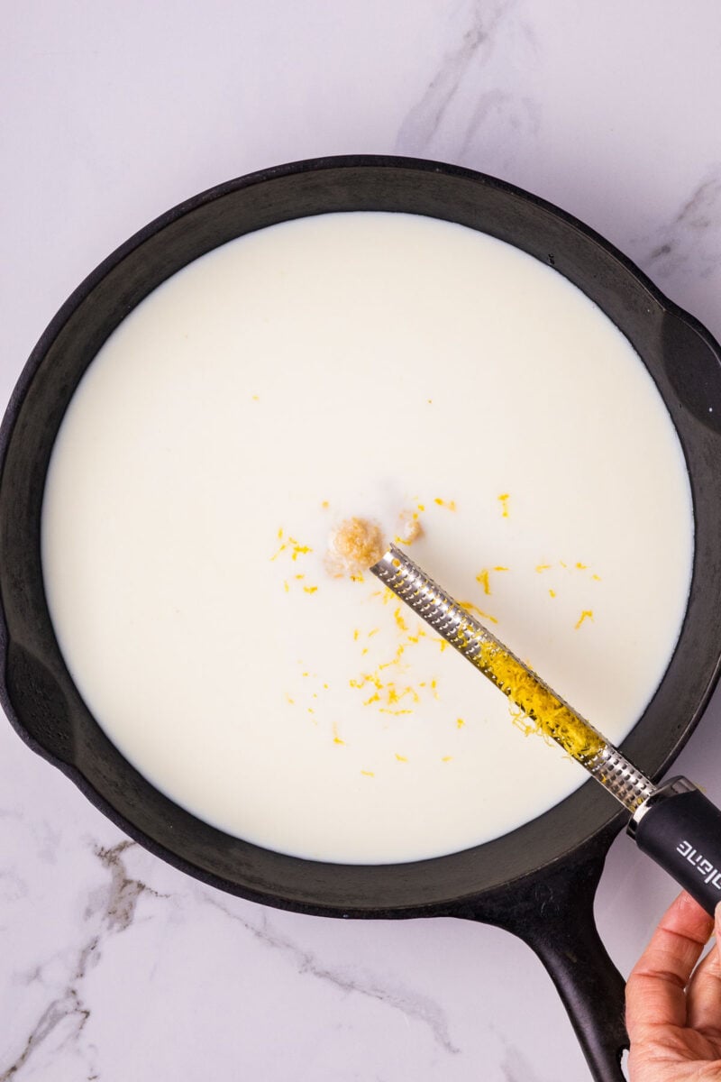 A hand grates lemon zest into a black skillet filled with milk, set on a white marble surface.