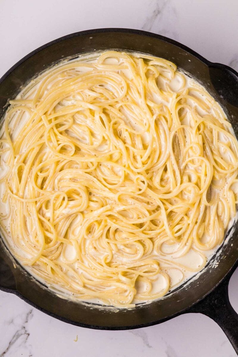 Linguini coated in a creamy white Alfredo sauce sits in a black skillet on a marble surface.