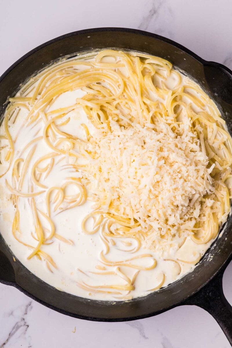 A skillet with cooked pasta in a creamy white alfredo sauce, topped with a mound of grated cheese, ready to be mixed together.