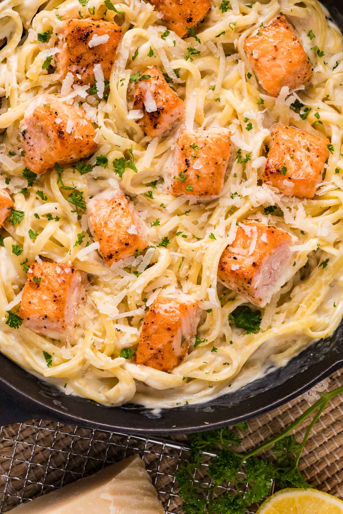 A close-up of creamy alfredo topped with salmon bites, grated cheese, and chopped parsley in a black dish.