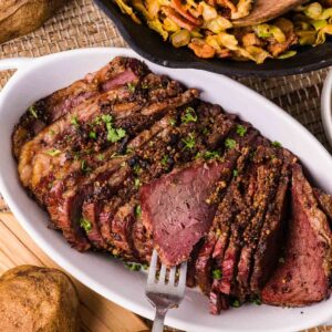 Sliced corned beef on a white platter with a fork, surrounded by baked potatoes and a skillet of cooked cabbage.