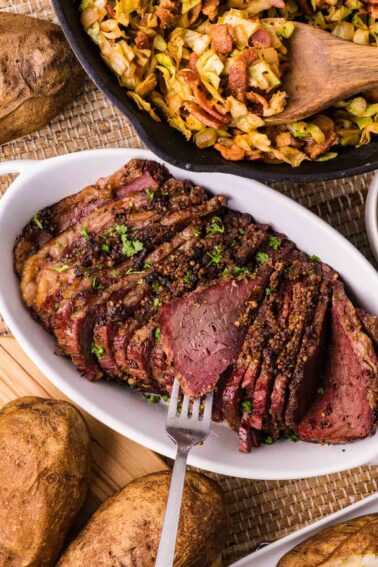 Sliced corned beef on a white platter with a fork, surrounded by baked potatoes and a skillet of cooked cabbage.