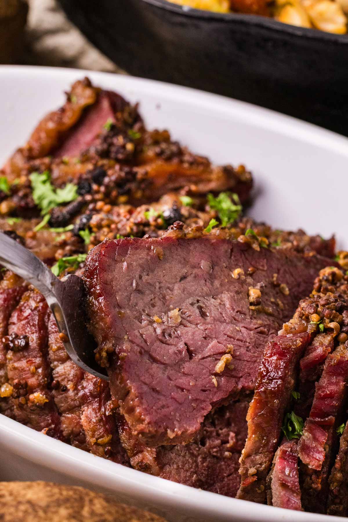 Sliced smoked and braised corned beef brisket garnished with herbs in a white dish, with a fork lifting one slice.