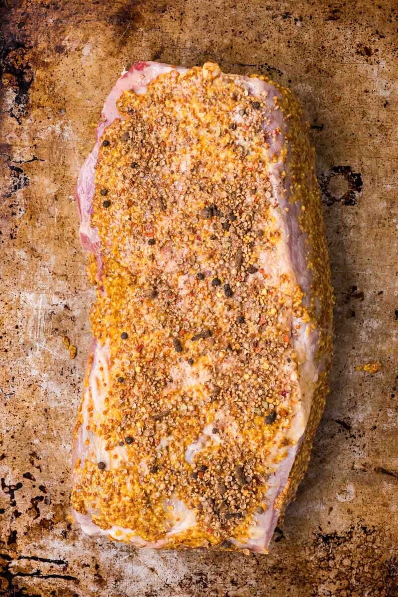 A raw corned beef coated with a coarse mustard and spice mixture rests on a worn metal baking sheet.
