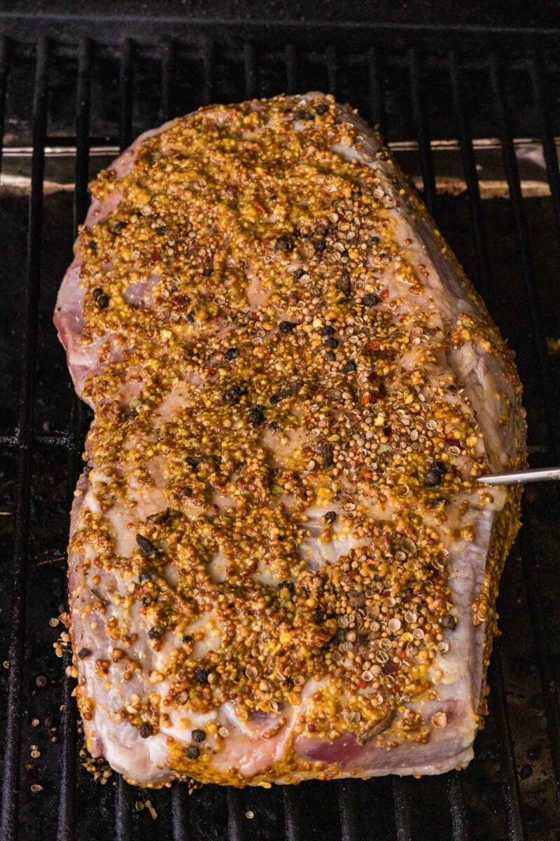 A corned beef coated with a coarse mustard and spice rub is being smoked on a grill with a meat thermometer inserted.