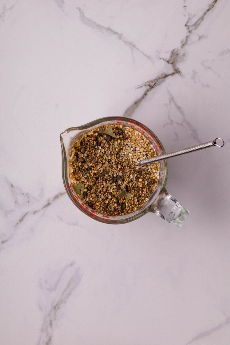 A glass measuring cup filled with mixed dry corned beef pickling spices and a metal spoon, placed on a white marble surface.