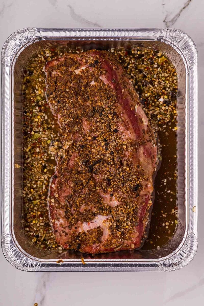 A seasoned smoked corned beef brisket sits in a foil tray, with braising liquid and pickling spices.