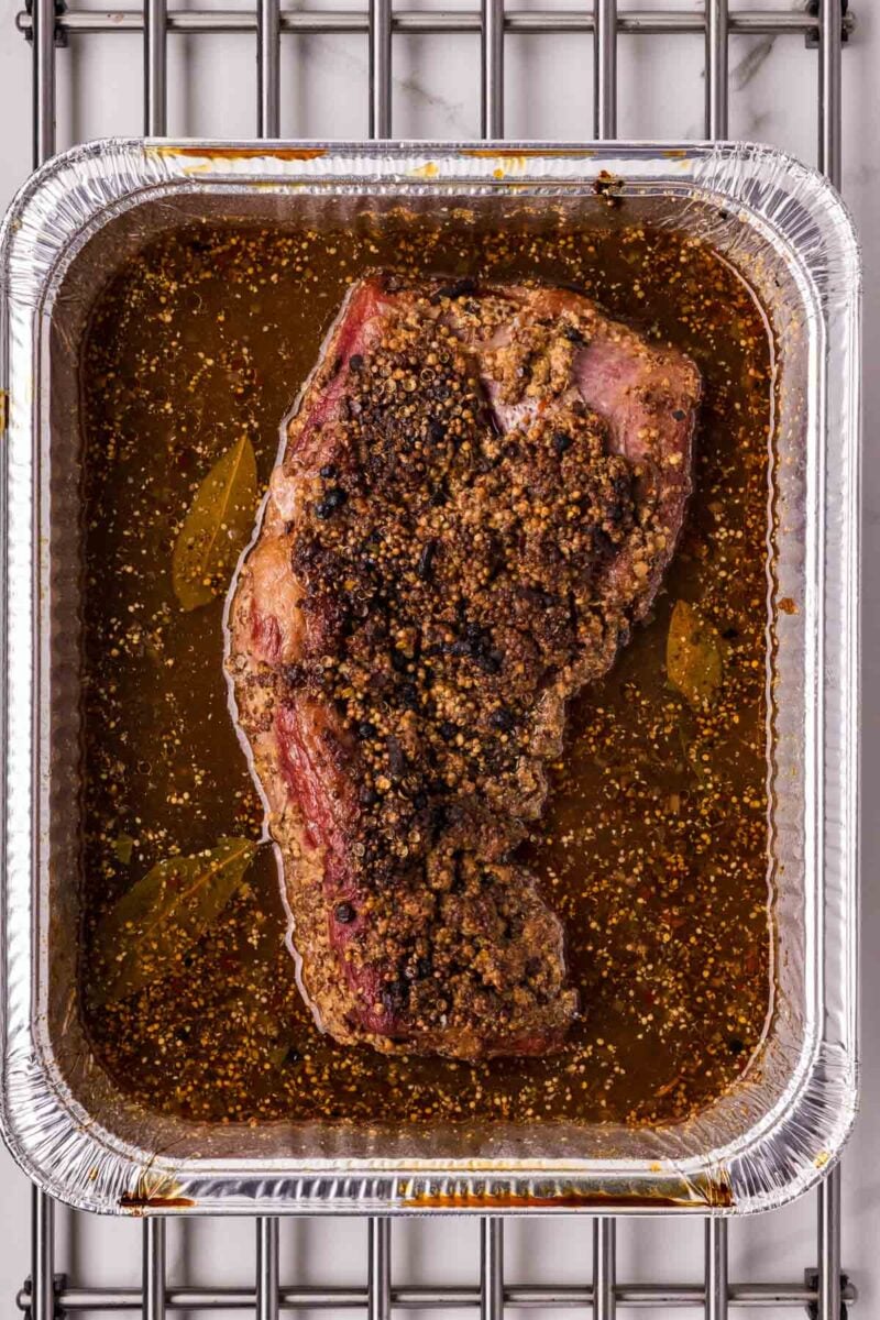 A cooked, seasoned smoked corned beef of meat sits in braising liquid inside a rectangular aluminum tray on a metal rack.