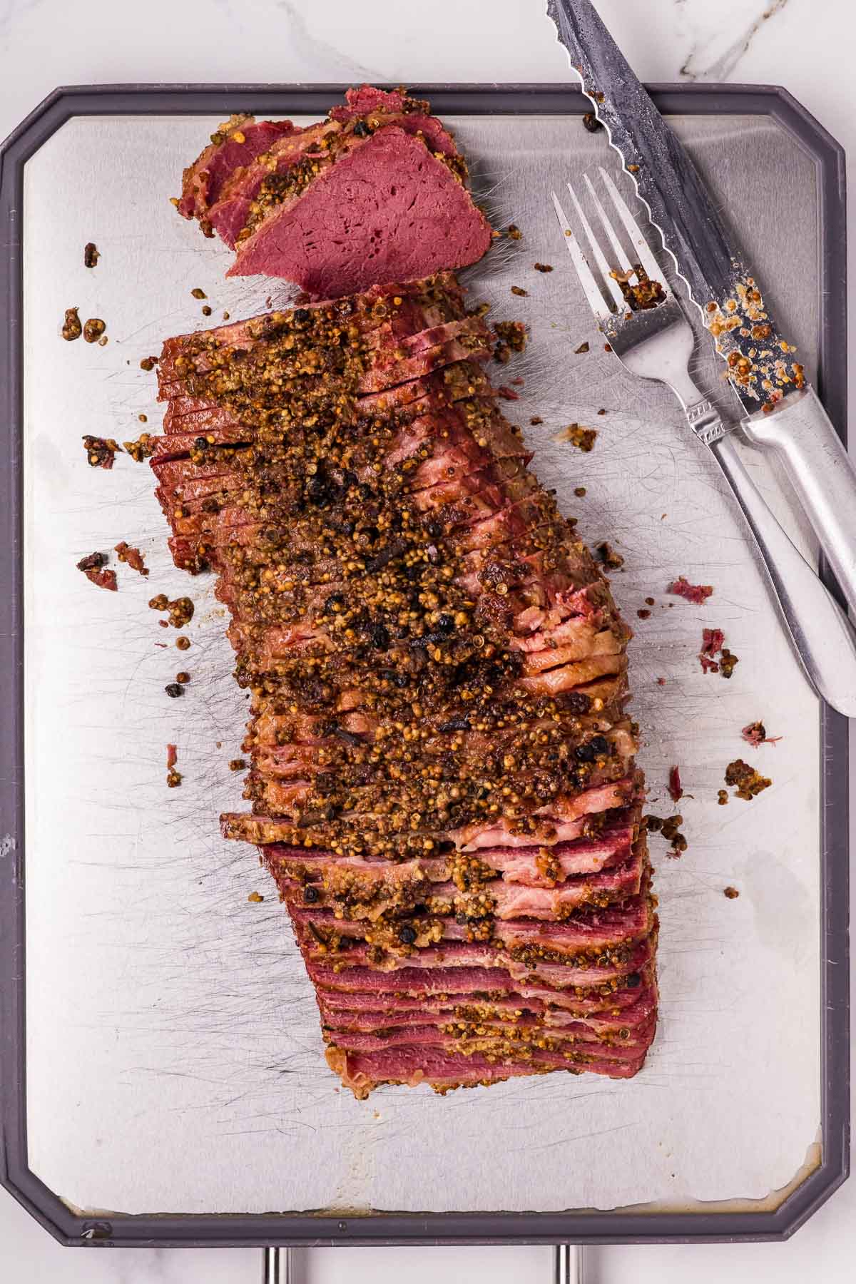 Sliced corned beef with a mustard and spice crust on a cutting board, accompanied by a knife and fork.
