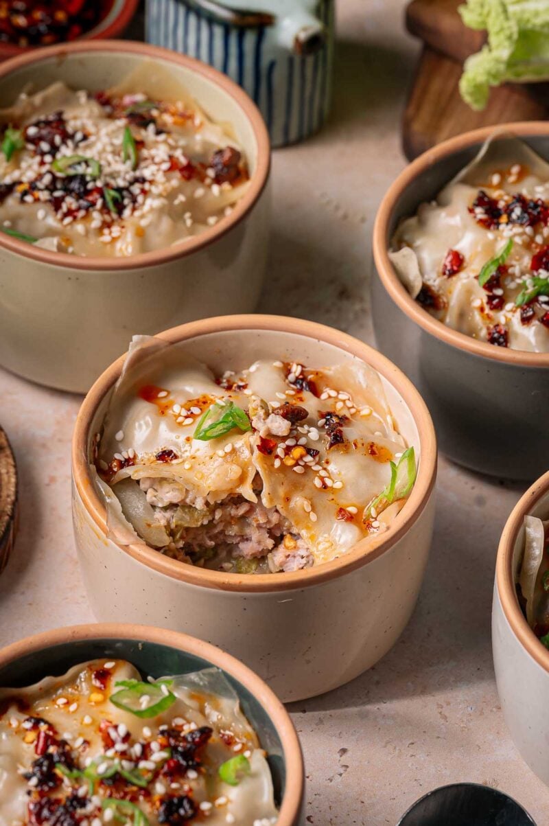 Individual ceramic bowls filled with dumpling lasagna topped with chili oil, sliced scallions, and sesame seeds, arranged on a light-colored surface.