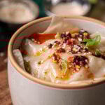 A bowl of dumpling lasagna topped with chili oil, sesame seeds, and green onion slices, with small bowls of seasoning in the background.