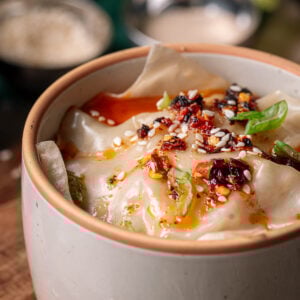 A bowl of dumpling lasagna topped with chili oil, sesame seeds, and green onion slices, with small bowls of seasoning in the background.
