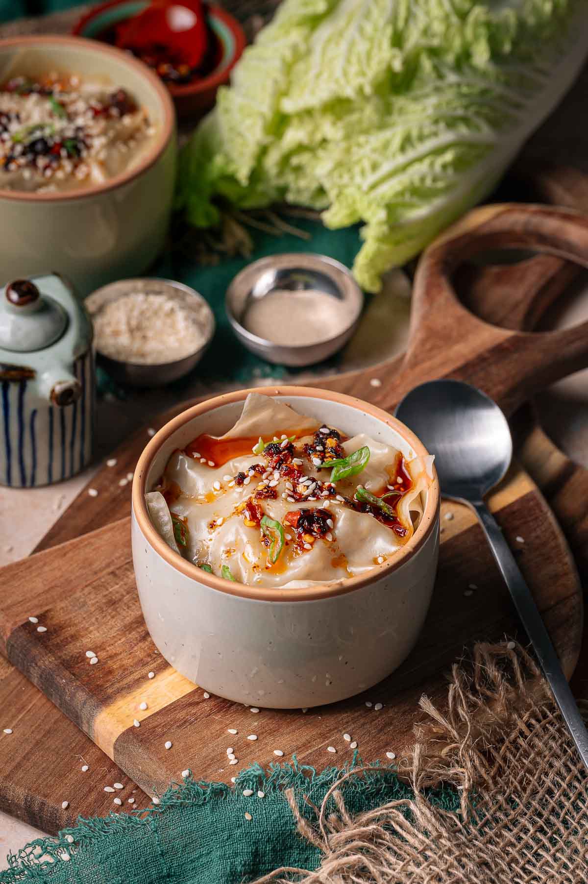 A ceramic bowl of dumpling lasagna topped with chili oil, green onions, and sesame seeds sits on a wooden board next to cabbage, sauces, and a spoon.