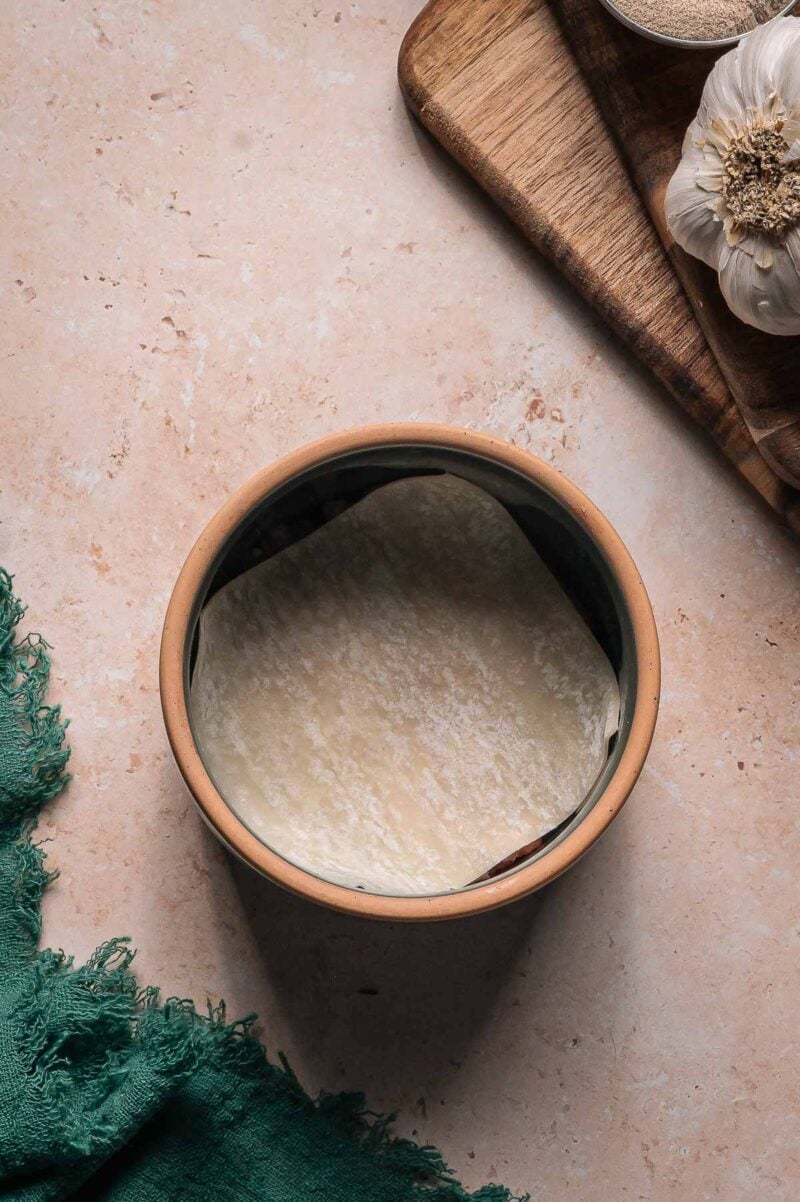 A ceramic bowl with a single dumpling wrapper arranged inside, placed on a beige countertop near a wooden board with a garlic bulb and a green cloth.