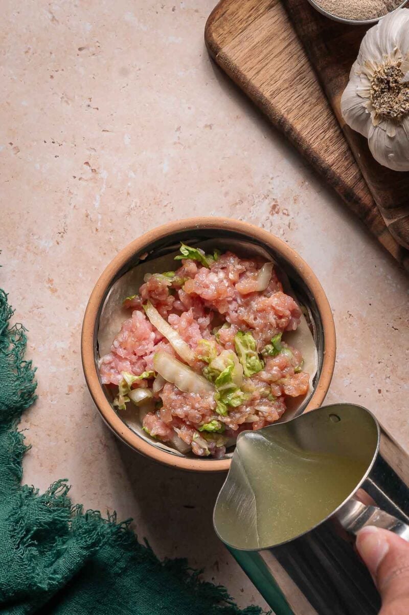 A ramekin of raw dumpling fillingsits on a counter as broth is poured in; garlic and a wooden board are nearby.