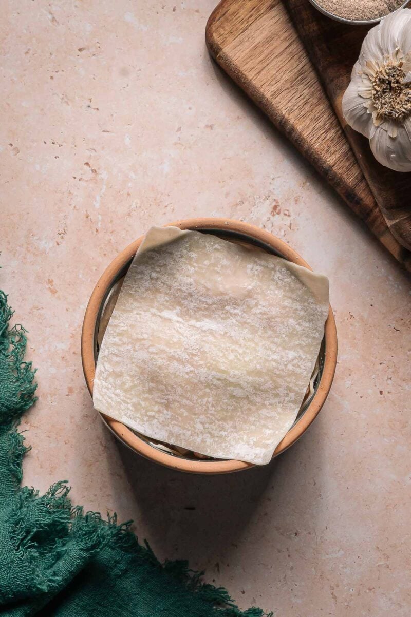 A single square sheet of uncooked dumpling wrapper layered over dumpling filling in a ramekin on a beige countertop, with a green cloth, a bulb of garlic, and a wooden board nearby.