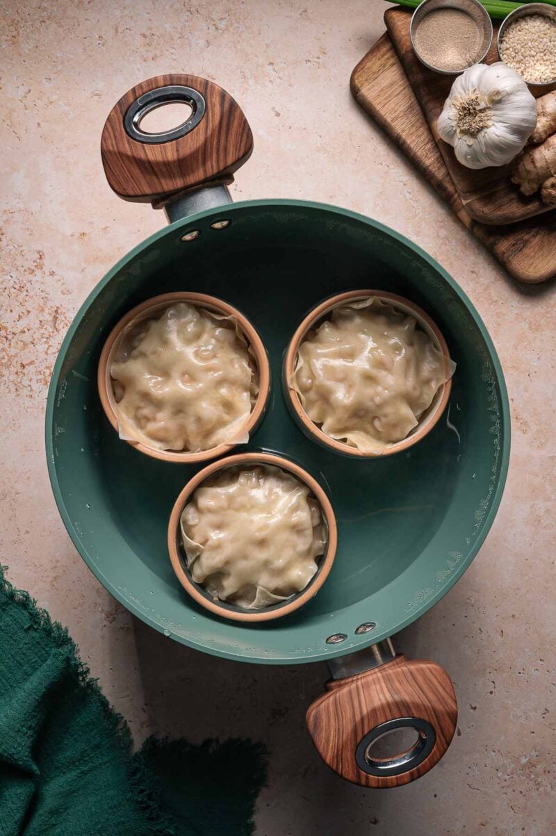 Three small bowls of dumpling lasagna are placed in a green pot of water, with garlic, ginger, and seasonings on a cutting board nearby.