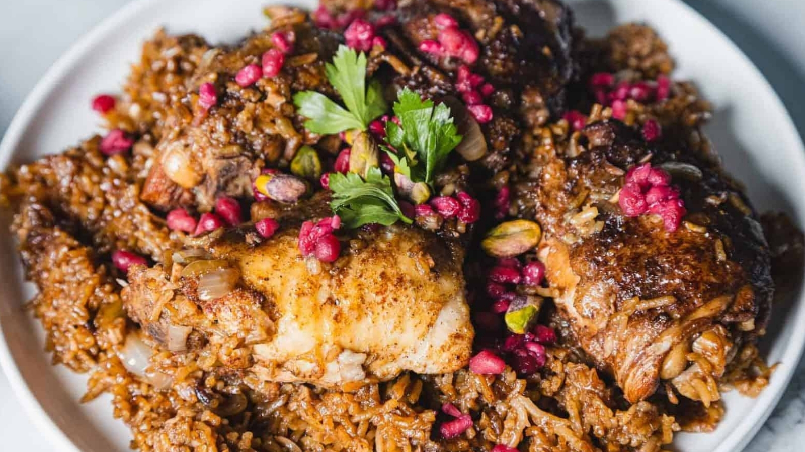 A close-up of a dish featuring seasoned rice topped with cooked chicken pieces. The meal is garnished with fresh parsley, pomegranate seeds, and pistachios, served on a white plate.