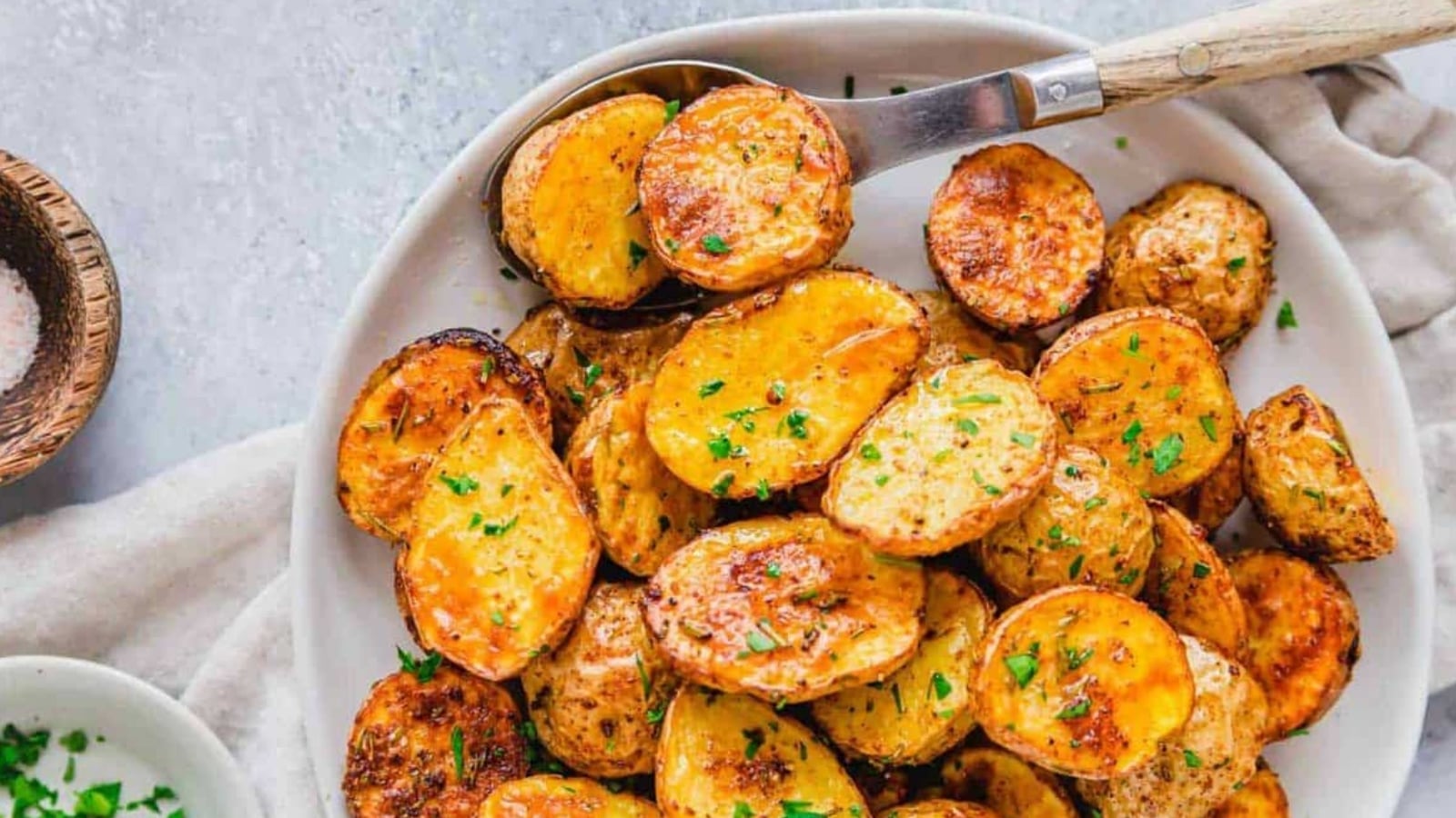 A white plate filled with roasted potato halves garnished with chopped herbs, with a serving spoon resting on the plate.