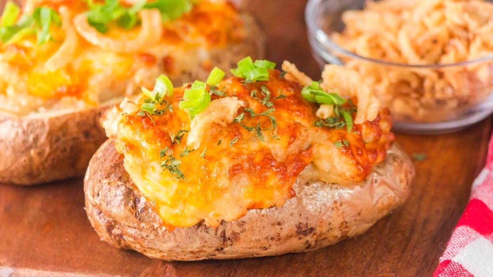 A baked potato topped with melted cheese, green onions, and crispy fried onions, with a bowl of fried onions in the background.