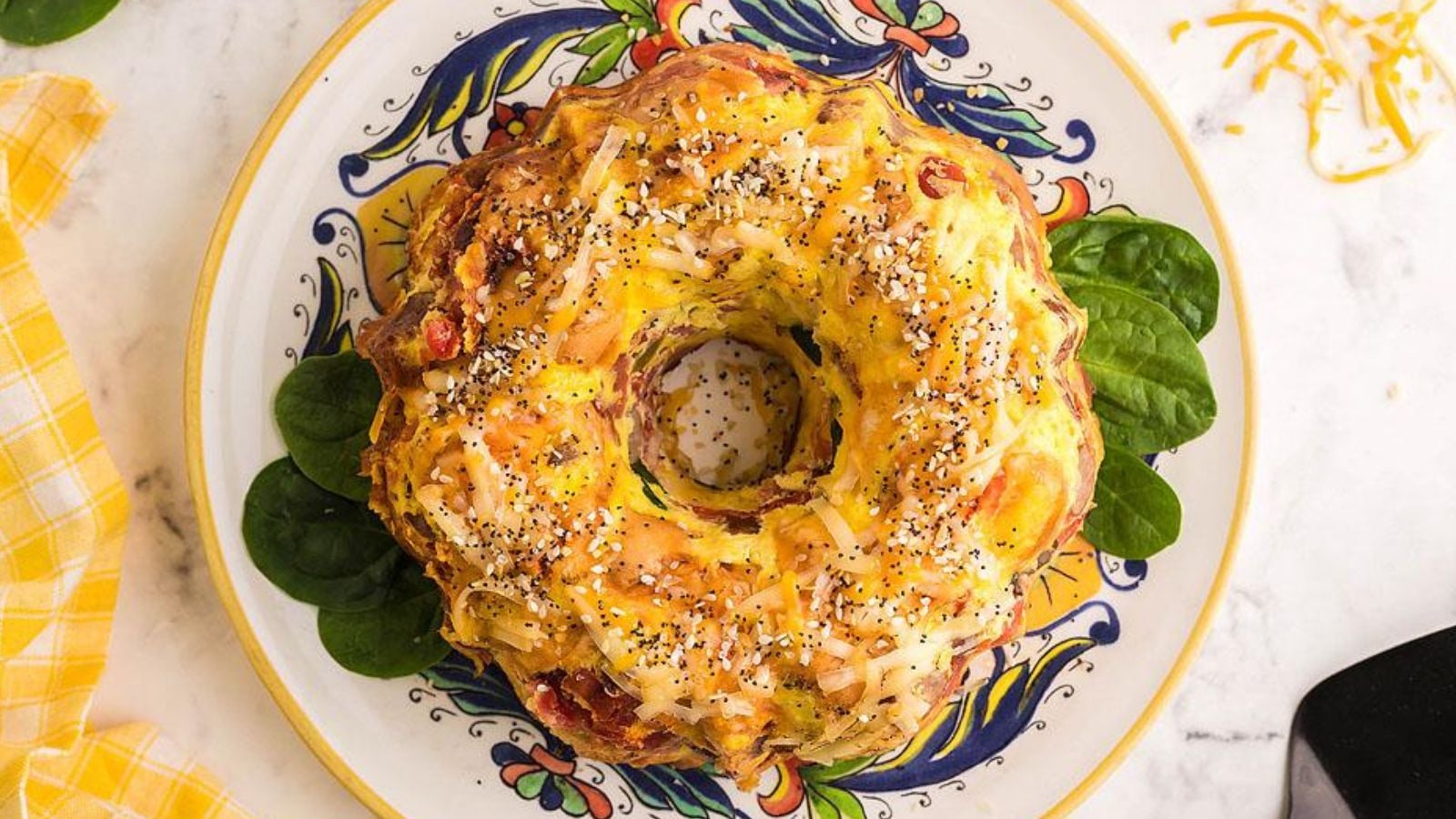 A savory bundt cake topped with shredded cheese and seeds sits on a decorative plate with spinach leaves arranged underneath.