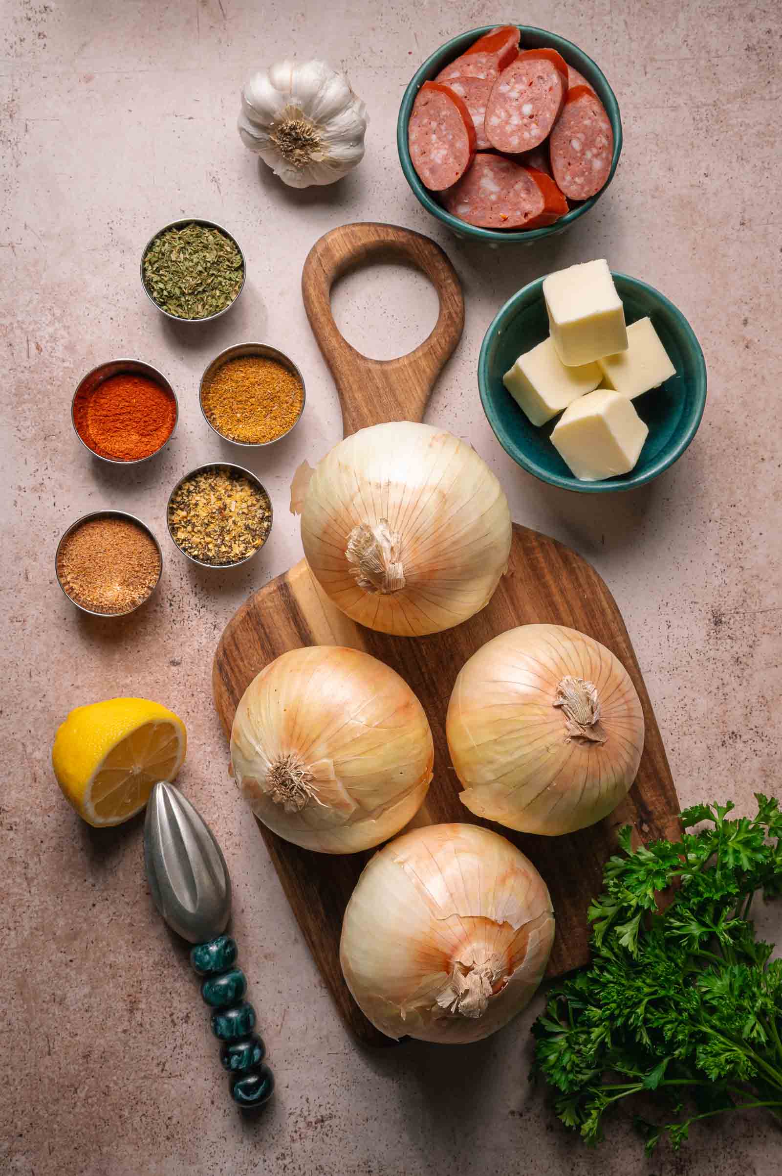 Overhead view of onions on a wooden board, surrounded by butter cubes, sausage slices, garlic, lemon, fresh parsley, and small bowls of assorted spices.