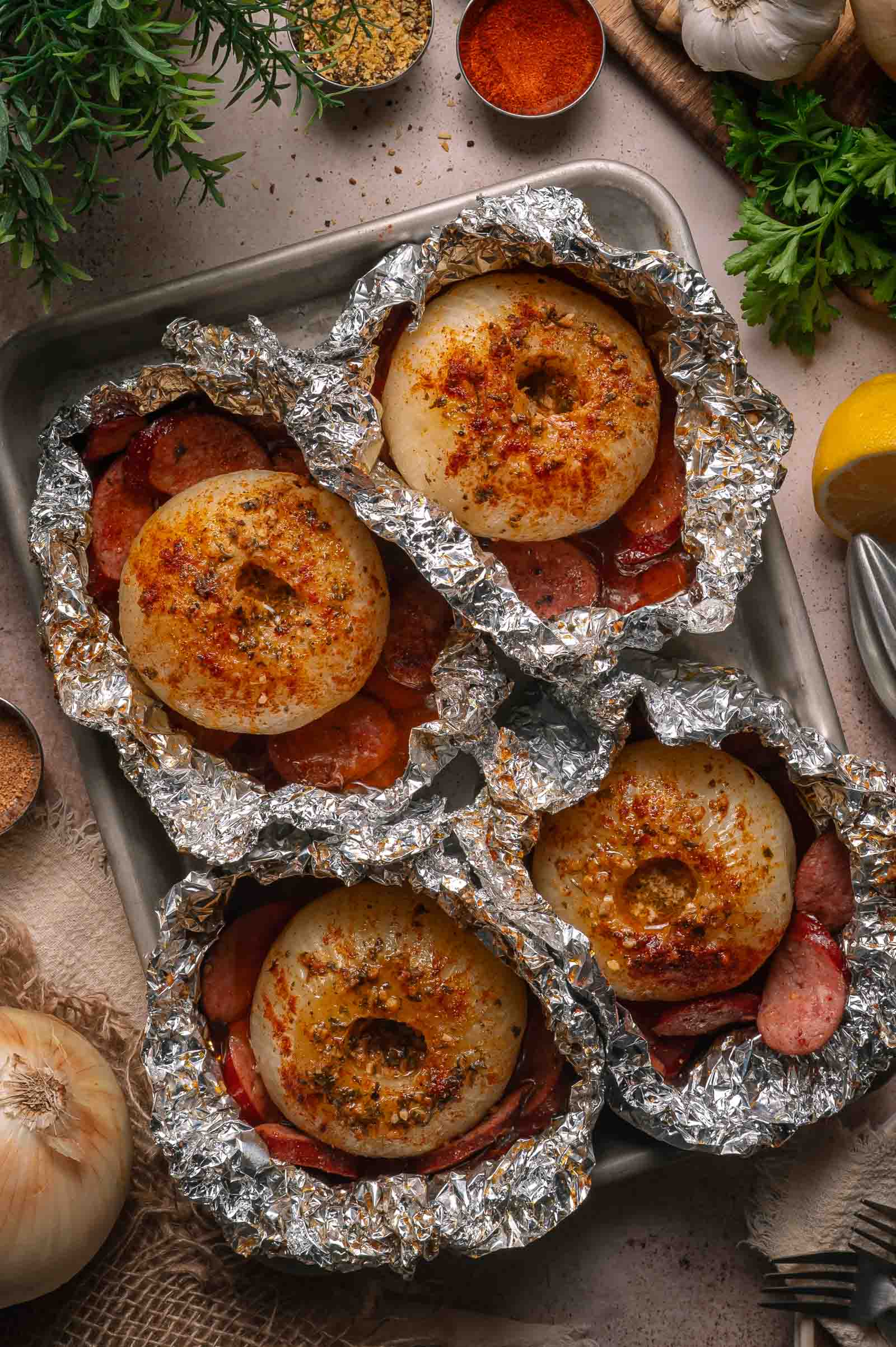 Four foil packets opened to expose a seasoned onion boil, sausage slices placed on a baking tray surrounded by herbs and spices.