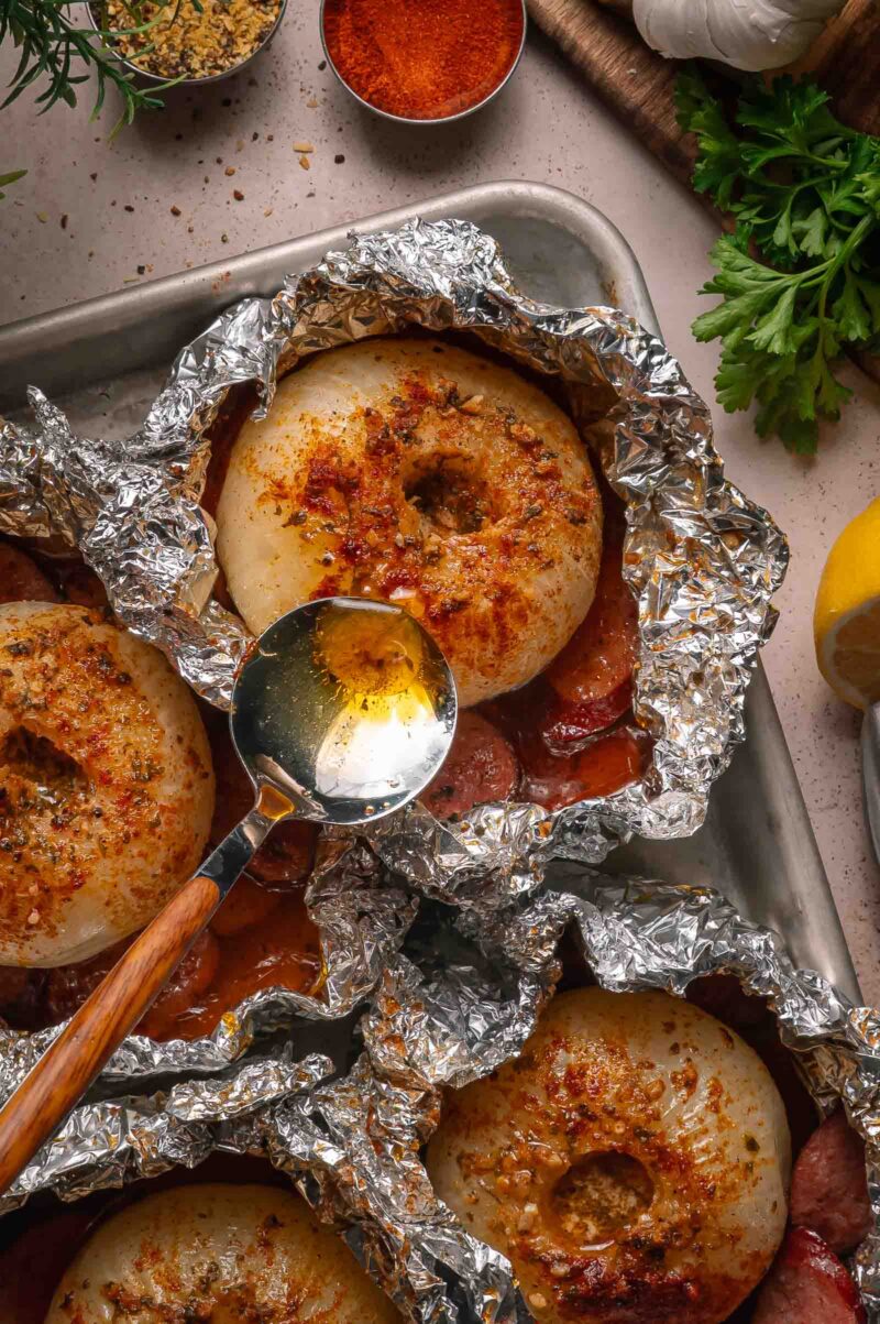 Boiled onions seasoned with spices being unwrapped from aluminum foil, and baked on a tray. A spoon drizzles liquid over one onion. Surrounding ingredients include parsley, garlic, and lemon.