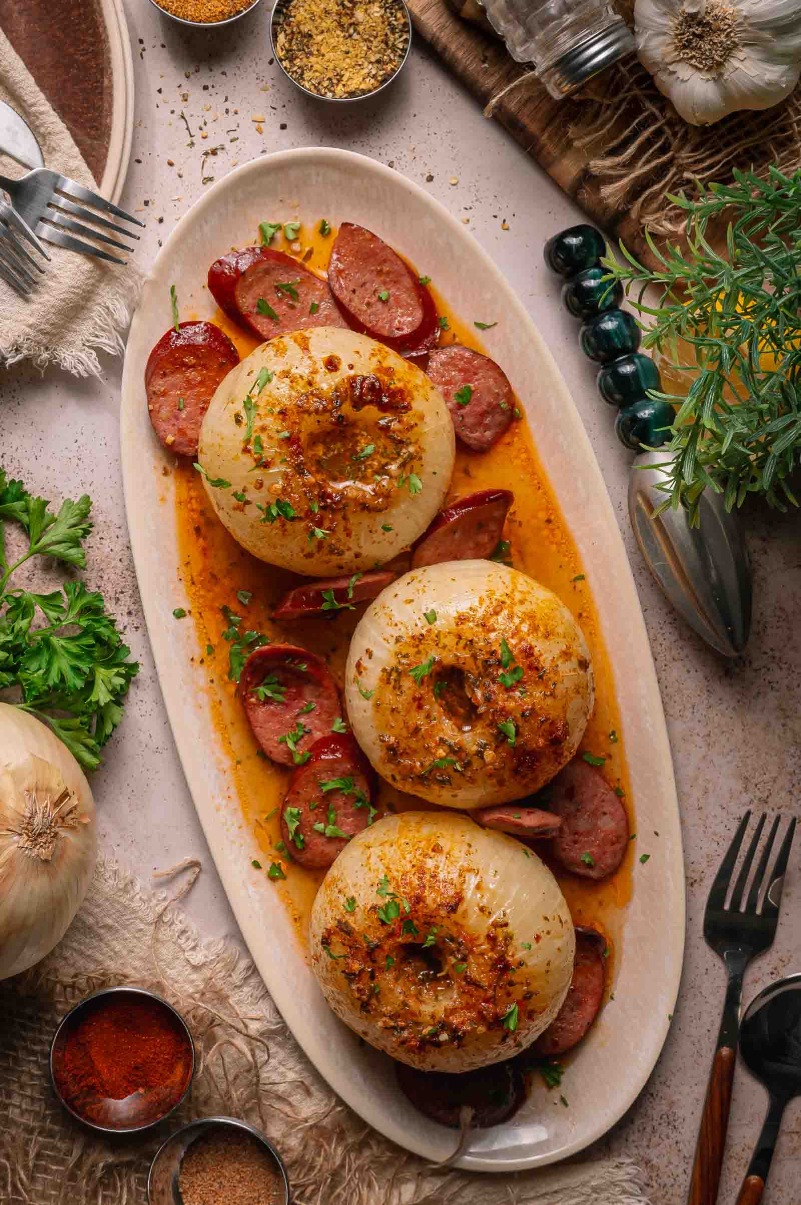 Three boiled onions served on an oval plate with sliced andouille and garnished with herbs, surrounded by seasonings and tableware.