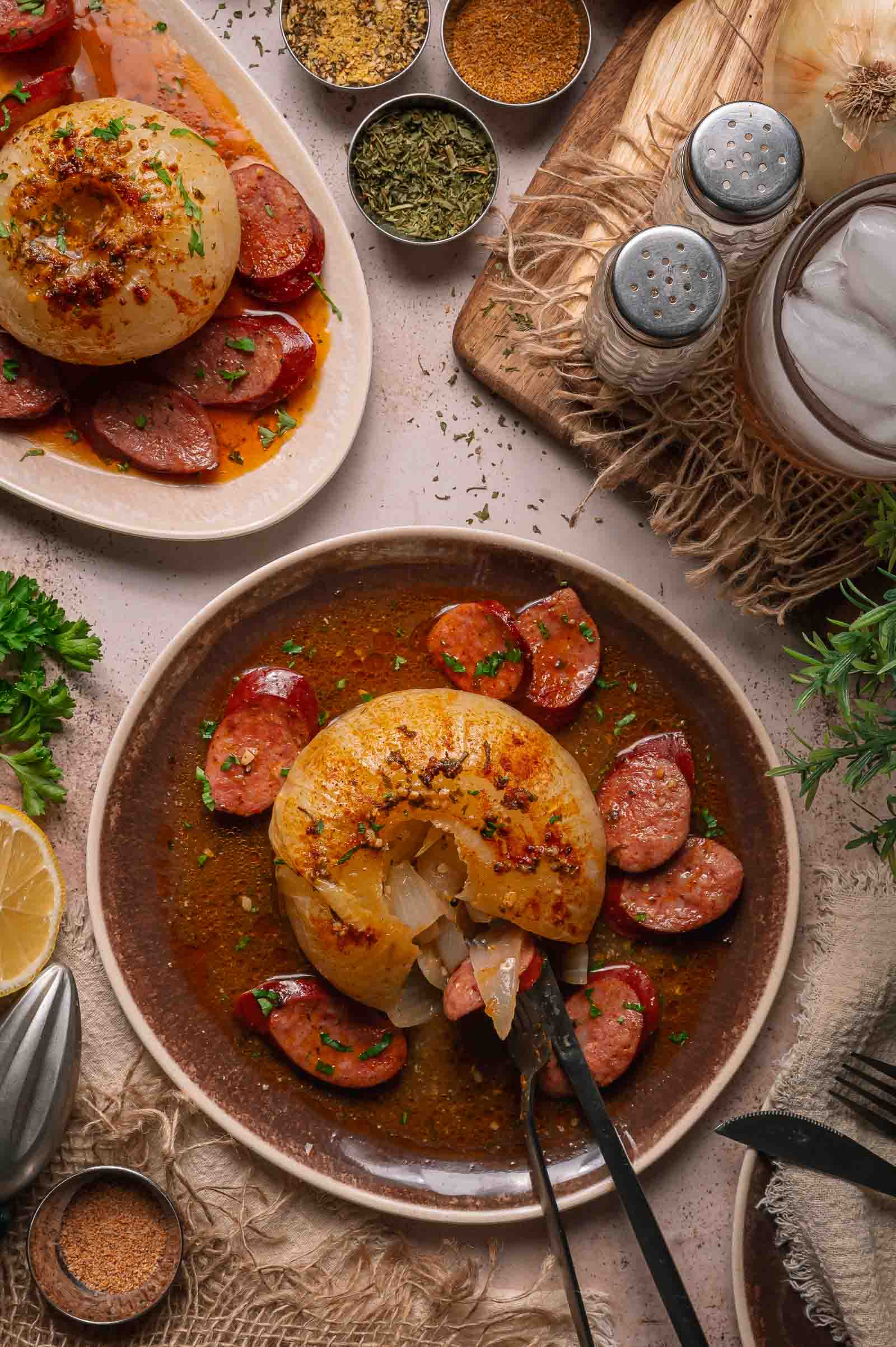 A plate with a boiled onion topped with herbs and spices, surrounded by sliced andouille in sauce, next to seasonings and a drink with ice.