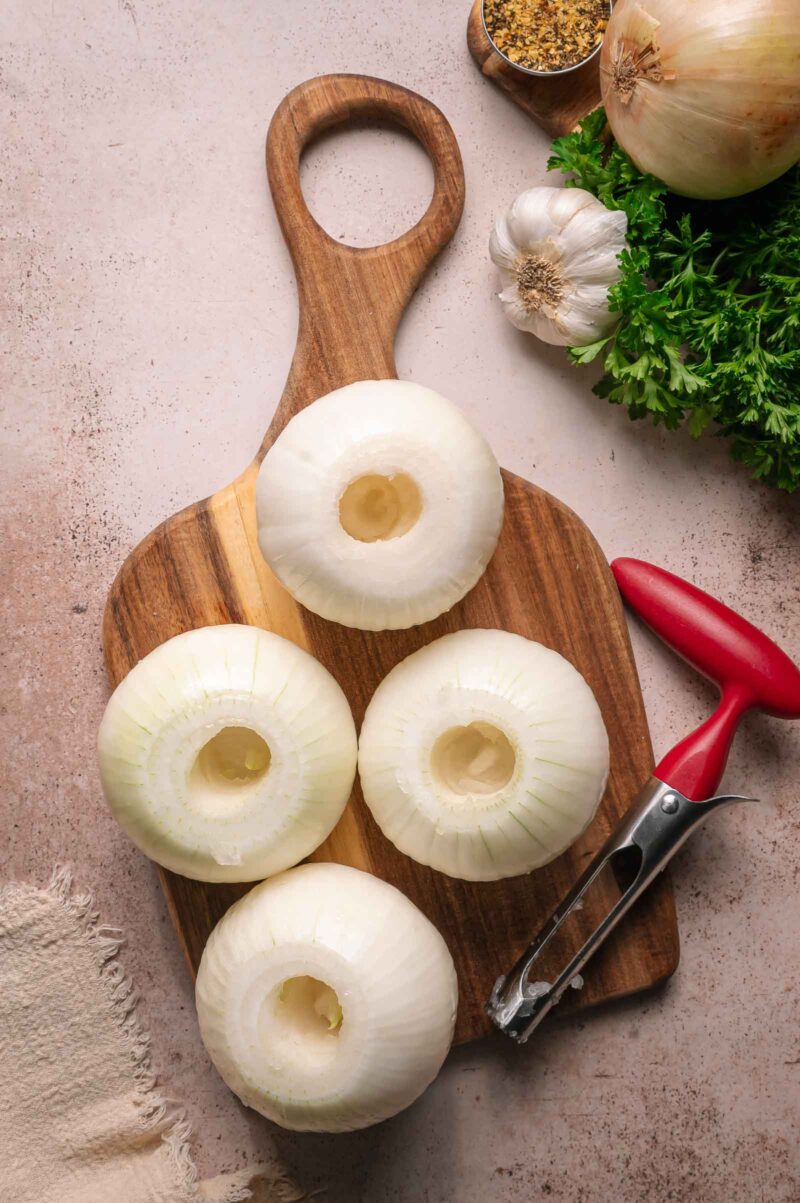 Four cored, peeled onions sit on a wooden cutting board next to a vegetable peeler, garlic, parsley, an onion, and a small bowl of seasoning.