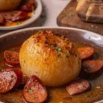 A whole onion served in buttery sauce with sliced andouille on a brown plate, garnished with chopped herbs.