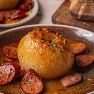 A whole onion served in buttery sauce with sliced andouille on a brown plate, garnished with chopped herbs.
