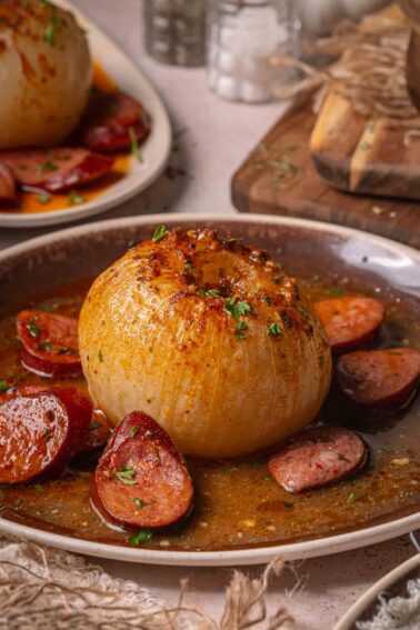 A whole onion served in buttery sauce with sliced andouille on a brown plate, garnished with chopped herbs.