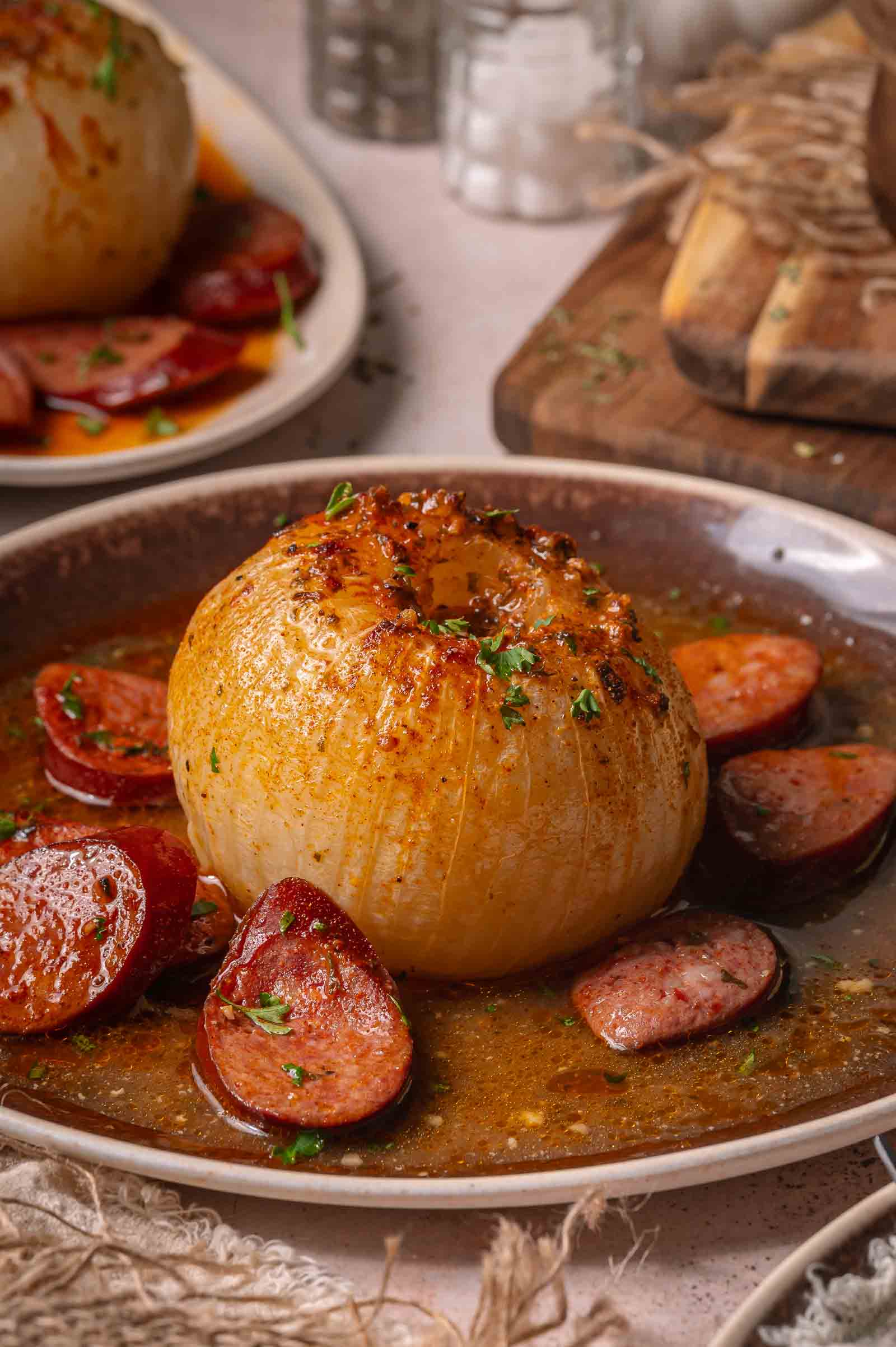 A whole onion served in buttery sauce with sliced andouille on a brown plate, garnished with chopped herbs.